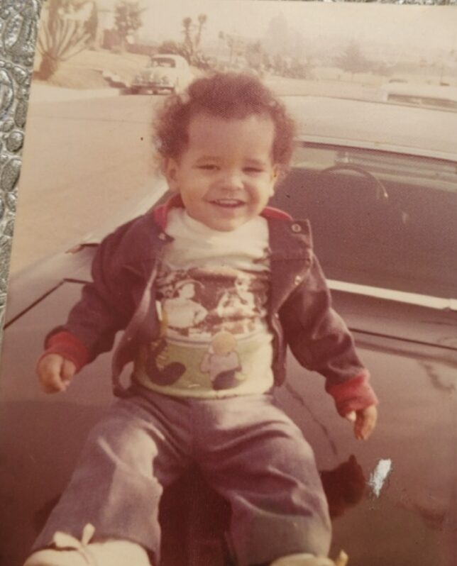 Nanon as a toddler, smiling, sitting on the hood of a car.