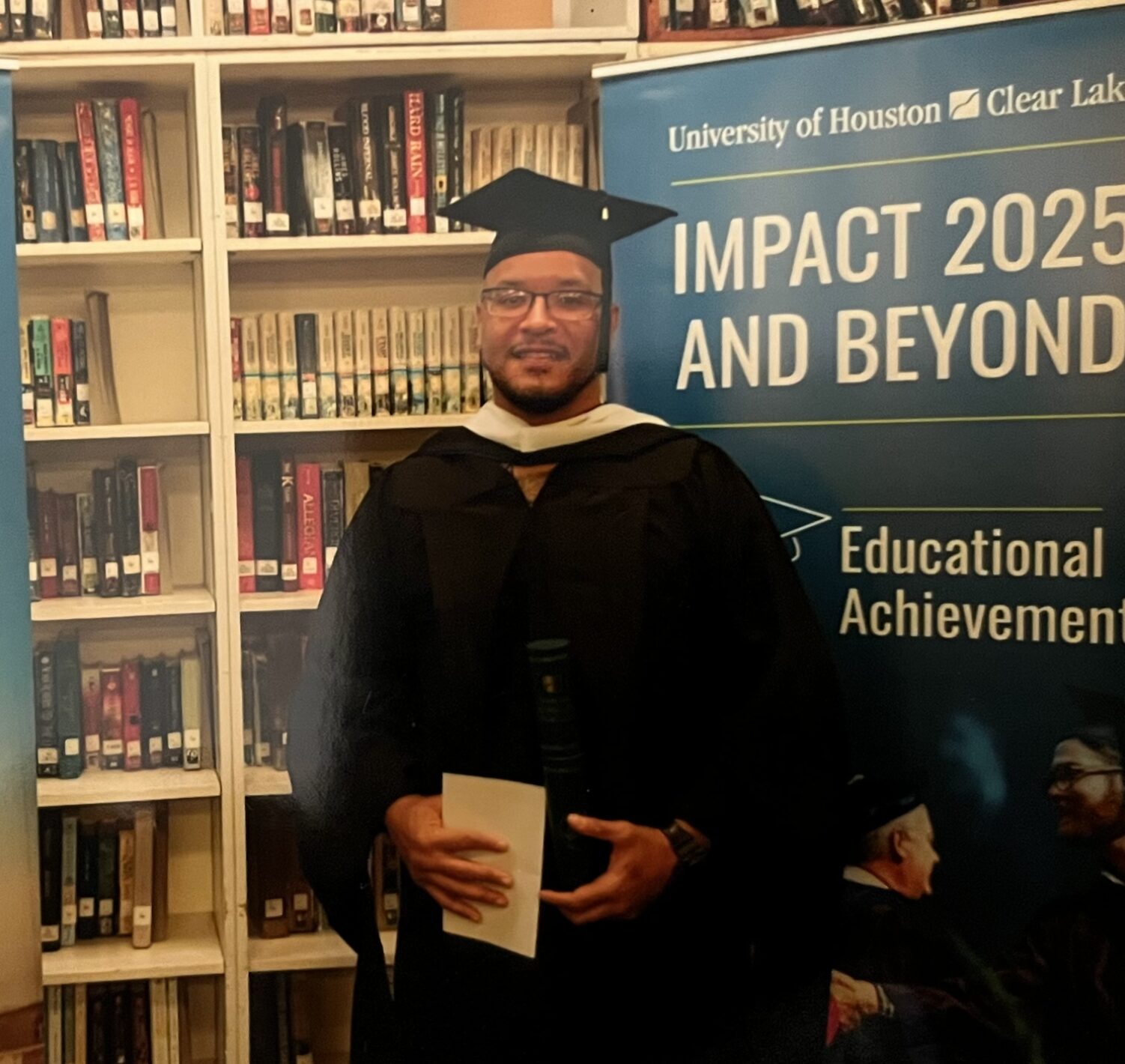 Nanon in cap and gown in front of bookshelves and a University of Houston-Clear Lake banner.